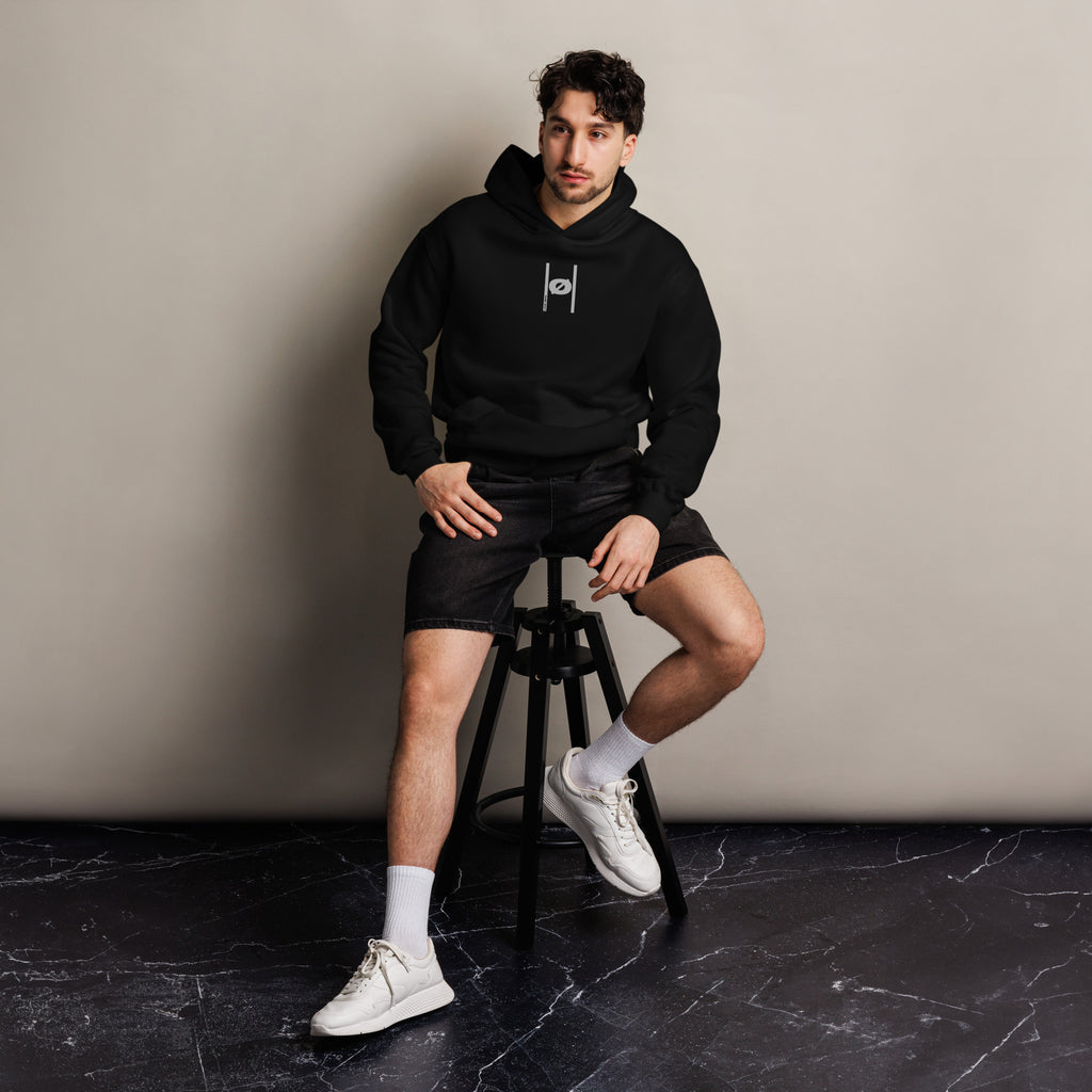 Man wearing a black hoodie and shorts sitting on a stool against a beige wall.